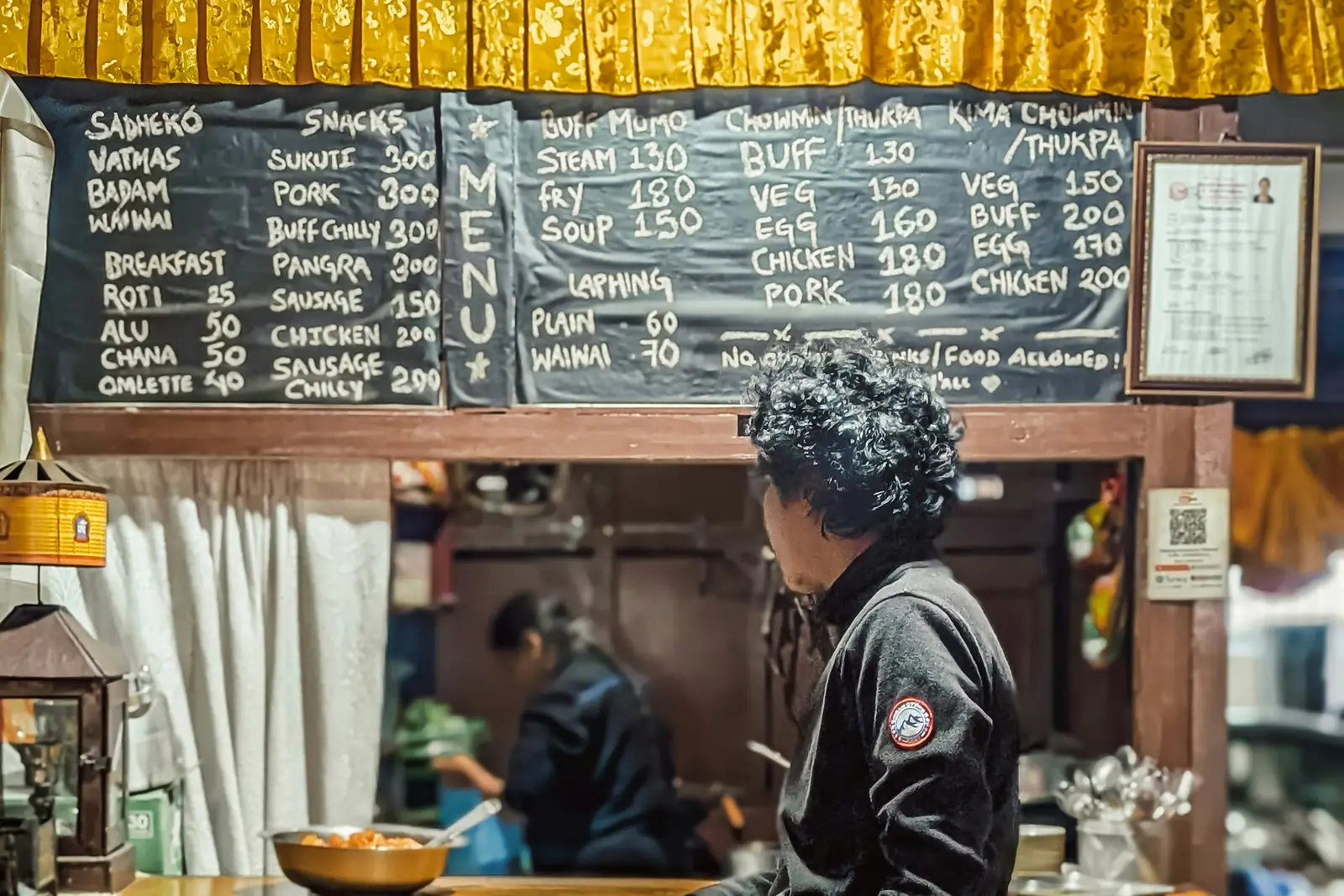 A customer waiting at the counter of a small restaurant in Chabahil, Kathmandu, under a chalkboard menu